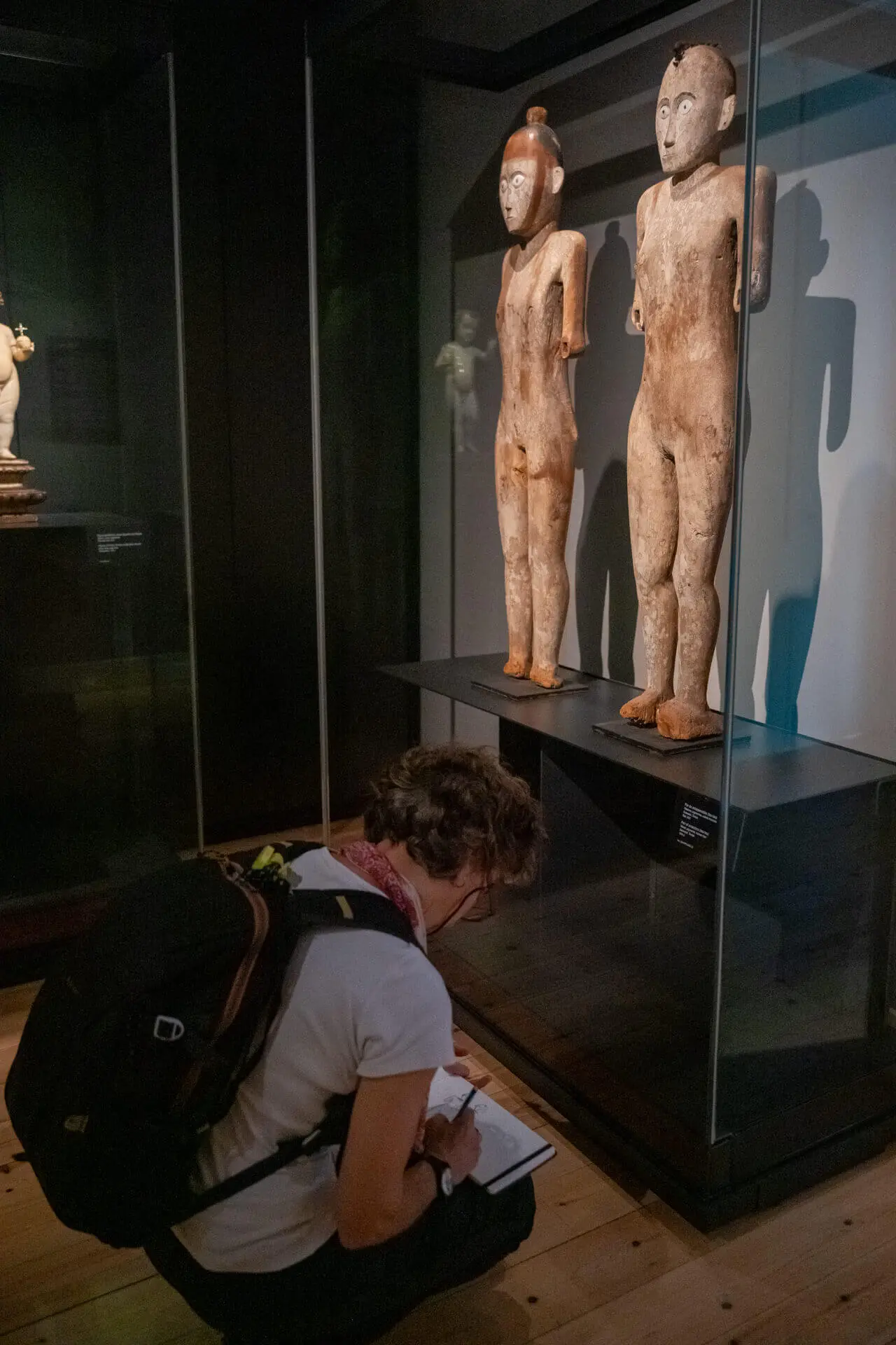 Ilustradora agachada a desenhar diante de duas esculturas de madeira em vitrine