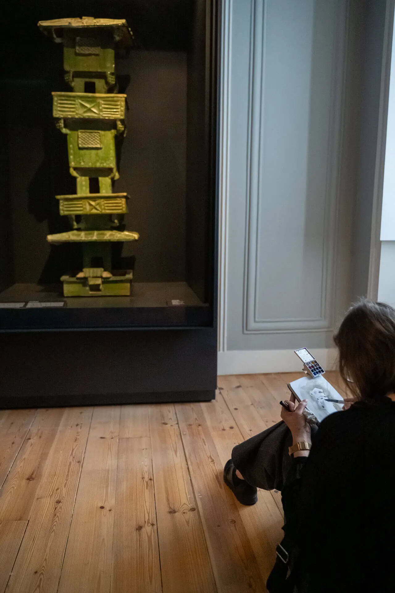 Ilustradora sentada no chão a desenhar diante de uma escultura alta, verde e amarela, em vitrine