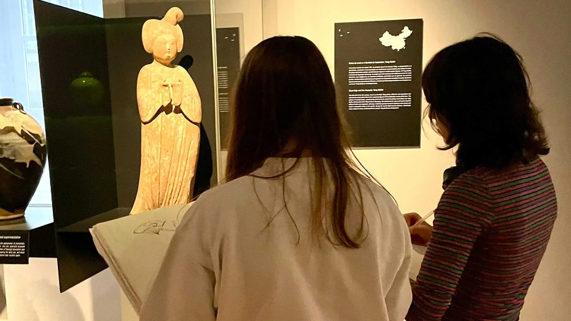 Duas alunas a desenhar diante de uma figura de terracota numa sala de exposição
